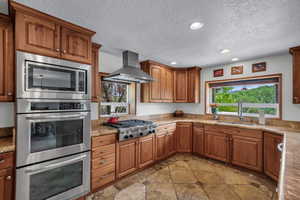 Kitchen featuring stainless steel appliances, wall chimney range hood, a textured ceiling, brown cabinets, and recessed lighting