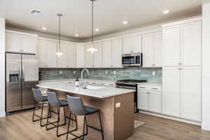 Kitchen with stainless steel appliances, light wood finished floors, a kitchen island with sink, light countertops, and a breakfast bar