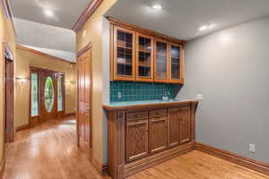 Bar featuring glass insert cabinets, tasteful backsplash, light wood-style flooring, crown molding, and brown cabinets