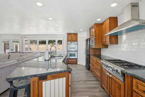 Kitchen with wall chimney range hood, open floor plan, stainless steel appliances, brown cabinets, and a breakfast bar area