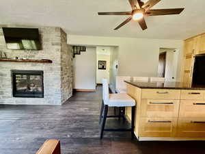 Kitchen featuring light brown cabinets, ceiling fan, dark stone countertops, dark wood finished floors, and a stone fireplace