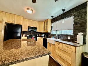 Kitchen featuring light brown cabinets, black appliances, tasteful backsplash, glass insert cabinets, and dark stone counters