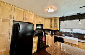 Kitchen featuring light brown cabinets, black appliances, tasteful backsplash, a textured ceiling, and glass insert cabinets
