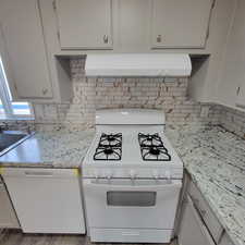 Kitchen with white appliances, exhaust hood, light stone countertops, and decorative backsplash