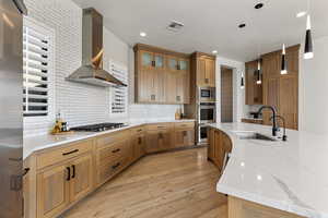 Kitchen with wall chimney exhaust hood, decorative backsplash, light wood-style floors, glass insert cabinets, and recessed lighting