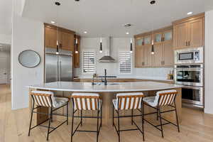 Kitchen with light wood finished floors, backsplash, built in appliances, a large island with sink, and brown cabinetry