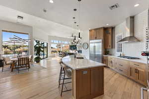 Kitchen with brown cabinets, wall chimney range hood, light wood-type flooring, a kitchen breakfast bar, and open floor plan