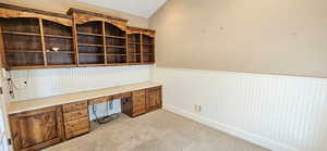 Unfurnished office featuring built in desk, wainscoting, light carpet, and lofted ceiling