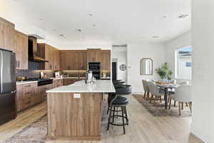 Kitchen featuring a center island with sink, stainless steel appliances, wall chimney exhaust hood, light wood-style floors, and recessed lighting