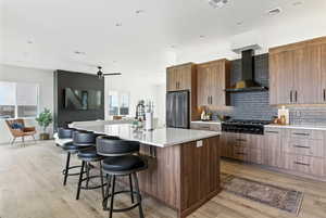 Kitchen featuring a kitchen bar, a center island with sink, wall chimney range hood, backsplash, and light countertops