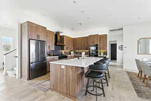 Kitchen featuring freestanding refrigerator, a center island with sink, wall chimney range hood, backsplash, and light countertops