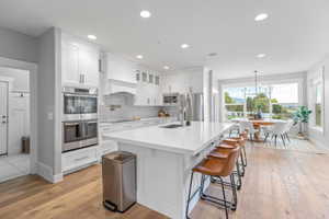 Kitchen featuring tasteful backsplash, stainless steel appliances, white cabinetry, light wood-type flooring, and a kitchen island with sink