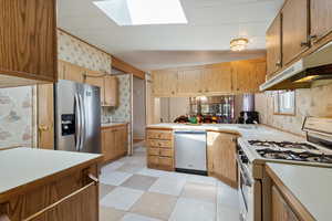 Kitchen featuring wallpapered walls, stainless steel appliances, a skylight, light countertops, and under cabinet range hood