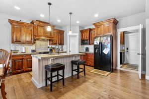 Kitchen with black appliances, light wood-type flooring, tasteful backsplash, a kitchen island with sink, and a breakfast bar