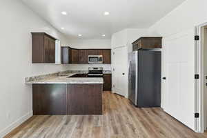 Kitchen with stainless steel appliances, dark brown cabinets, light wood-type flooring, a peninsula, and light stone countertops