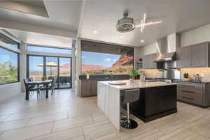 Kitchen featuring backsplash, an island with sink, wall chimney exhaust hood, open shelves, and dark stone countertops
