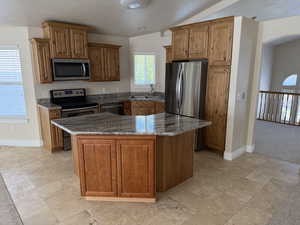 Kitchen featuring stainless steel appliances, dark stone counters, brown cabinetry, a kitchen island, and lofted ceiling