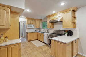 Kitchen featuring stainless steel appliances, under cabinet range hood, light countertops, light brown cabinets, and recessed lighting