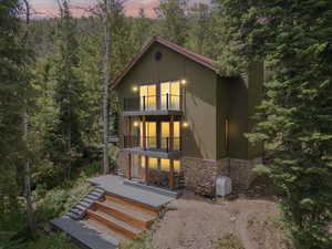 Front of house at dusk with a balcony, stone siding, a view of trees, and covered porch