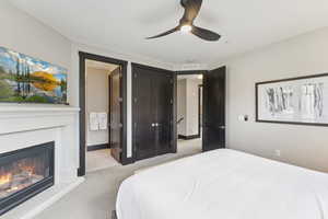 Bedroom featuring light colored carpet, a glass covered fireplace, ensuite bath, and a ceiling fan