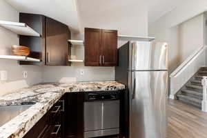Kitchen featuring open shelves, stainless steel appliances, dark brown cabinets, light wood finished floors, and light stone counters