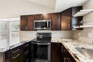 Kitchen with appliances with stainless steel finishes, dark brown cabinets, a peninsula, vaulted ceiling, and light stone counters