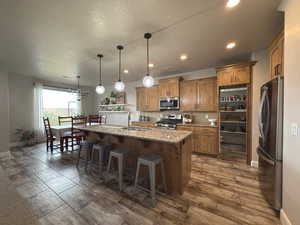 Kitchen featuring brown cabinets, stainless steel appliances, a kitchen bar, light stone counters, and a kitchen island with sink