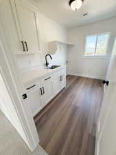 Utility room featuring wood-finish flooring, white cabinetry with black hardware, a stainless steel sink with a matte black faucet, a quartz-look countertop, and a window