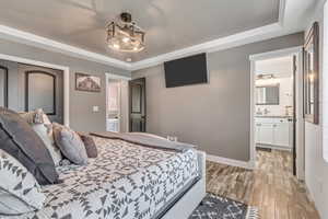 Bedroom with light wood-style flooring, a raised ceiling, ensuite bath, and crown molding
