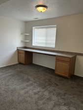 Unfurnished office with a textured ceiling, dark carpet, and built in desk