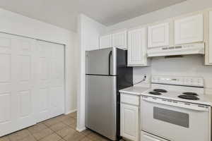 Kitchen with white electric stove, freestanding refrigerator, under cabinet range hood, light countertops, and light tile patterned floors