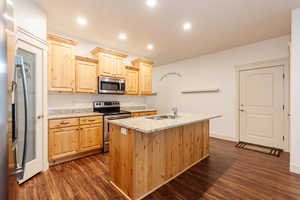 Kitchen with light brown cabinets, appliances with stainless steel finishes, dark wood finished floors, light stone counters, and a center island with sink