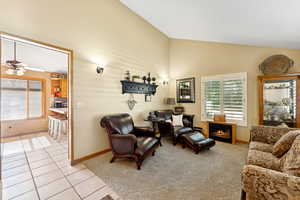 Living area with light colored carpet, ceiling fan, light tile patterned flooring, high vaulted ceiling, and a warm lit fireplace