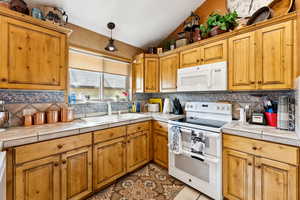 Kitchen with white appliances, vaulted ceiling, tile counters, tasteful backsplash, and brown cabinets