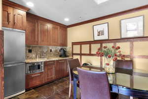 Kitchen featuring high end refrigerator, decorative backsplash, light stone countertops, ornamental molding, and brown cabinets