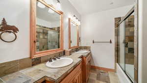 Full bath with double vanity, bath / shower combo with glass door, and stone finish flooring
