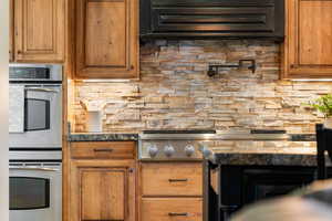 Kitchen with stainless steel appliances, brown cabinetry, backsplash, and range hood