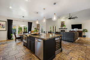 Kitchen with hanging light fixtures, appliances with stainless steel finishes, a glass covered fireplace, dark stone counters, and an island with sink