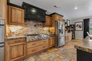 Kitchen featuring tasteful backsplash, stainless steel appliances, brown cabinetry, wall chimney range hood, and stone tile flooring