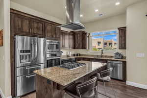 Kitchen featuring stainless steel appliances, dark brown cabinets, island range hood, a kitchen island, and light stone counters