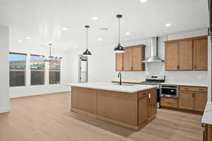 Kitchen featuring backsplash, stainless steel range with gas cooktop, light wood-style floors, wall chimney exhaust hood, and brown cabinetry