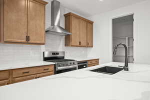 Kitchen featuring gas stove, wall chimney exhaust hood, decorative backsplash, brown cabinetry, and light stone countertops