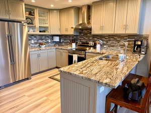 Kitchen featuring a peninsula, appliances with stainless steel finishes, wall chimney exhaust hood, a kitchen bar, and backsplash