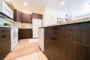 Kitchen featuring white appliances, dark brown cabinets, light wood finished floors, recessed lighting, and light stone counters
