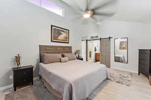 Bedroom featuring light wood-style floors, a barn door, high vaulted ceiling, ceiling fan, and connected bathroom
