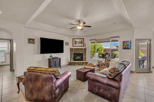 Living area featuring light tile patterned floors, a raised ceiling, a ceiling fan, arched walkways, and a stone fireplace