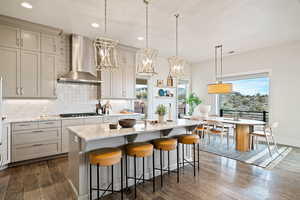 Kitchen featuring backsplash, wall chimney range hood, a kitchen island with sink, decorative light fixtures, and light stone counters