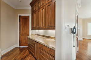 Kitchen with ornamental molding, dark wood-type flooring, light stone countertops, brown cabinetry, and recessed lighting