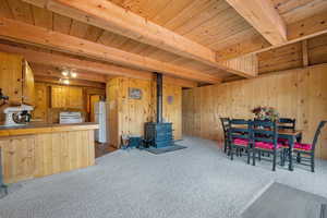 Dining area with a wood stove, a wooden ceiling with exposed beams, wooden walls, and carpet