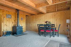 Carpeted dining room featuring wood walls, a wood stove, and a wooden ceiling with exposed beams